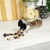 A spice grinder lying on its side with peppercorns spilled on a white wooden surface. In the background, there is a cutting board and a jar of pasta. Produktbild-Vorschau