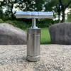 A metal pollen press tool sits on a textured stone surface, with blurred trees and rocks in the background. Produktbild-Vorschau