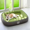 A corgi dog is lying on a green and gray dog bed in front of a window with green foliage visible outside. Produktbild-Vorschau