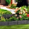 A garden bed with a stone-look plastic border, featuring various flowers and greenery, with a house and walkway in the background. Produktbild-Vorschau