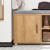 A close-up view of a wooden shoe cabinet with a gray upholstered seat on top. A brown hat and a brown boot are visible on the seat and in an open shelf respectively. Produktbild-Vorschau