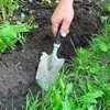 A hand holding a trowel is digging into the soil next to a garden bed with green plants and grass. Náhľad obrázku produktu