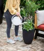 A person watering a plant in a black planter on a brick patio. Produktbild-Vorschau