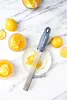 A Microplane zester is shown on a plate with lemon zest, surrounded by lemons and drinks, on a marble surface. Náhled obrázku produktu
