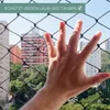 A hand is shown reaching through a black net, with buildings and trees visible in the background. Produktbild-Vorschau