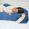 A woman is lying on a bed, resting her head on a large blue U-shaped pillow. Náhľad obrázku produktu