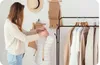 A woman is measuring a mannequin in a sewing studio, with sewing patterns and clothes on a rack in the background. Podgląd zdjęcia produktu