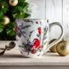 A large mug with a Christmas bird design sits on a surface next to a spoon and Christmas ornaments, with a decorated Christmas tree in the background. Voir les images du produit