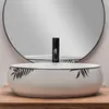 A white oval basin with a black leaf pattern sits on a wooden surface in front of a large round mirror. Náhled obrázku produktu