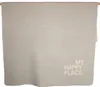 A light gray blanket with text 'MY HAPPY PLACE' printed on the bottom right corner, hanging from a wooden rod. Produktbild-Vorschau