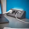 A gray power strip with multiple outlets and USB ports is plugged into a laptop on a desk, with a blue wall in the background. Produktbild-Vorschau