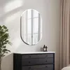 A frameless oval mirror is mounted on a wall above a dark dresser. A plant is to the left of the mirror, and curtains are to the right. Podgląd zdjęcia produktu