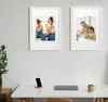 Two framed pictures of families hanging on a wall above a desk with a laptop, phone, and mug. Produktbild-Vorschau