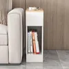 A narrow white nightstand with a wood-effect top sits next to a cream-colored sofa. The nightstand has a drawer at the top and an open shelf below, which holds four books. The background is a wood-paneled wall, and the floor is tiled in gray. Náhled obrázku produktu