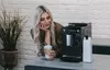 A woman is holding a latte next to a coffee machine. Produktbild-Vorschau