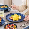 A person is eating a meal from a blue ceramic plate with spaghetti, chicken, and broccoli. Other dishes and a glass of water are also visible. Voir les images du produit