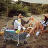 A couple is enjoying a picnic outdoors, toasting with drinks. A foldable wagon filled with picnic items is in the foreground. Produktbild-Vorschau