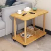 A small wooden side table with two shelves is placed next to a light gray sofa in a living room setting. The table has a vase with branches, a tissue box, a remote control, and a phone on the top shelf. The bottom shelf holds a basket with books and magazi Produktbild-Vorschau
