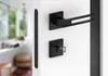 A modern black door handle set with a keyhole escutcheon is mounted on a white door. The background shows a blurred interior scene with a window and furniture. Produktbild-Vorschau