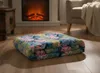 A folded blanket with a tropical leaf pattern is placed on a wooden floor in front of a fireplace and a chair. Podgląd zdjęcia produktu