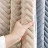 A hand is shown touching a beige, plush, quilted fabric with a chevron pattern. Behind it, a similar fabric in a lighter beige and a gray quilted fabric are visible. Náhled obrázku produktu