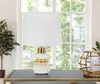 A table lamp with a white shade and a white and gold base sits on a dark surface in front of a window. Náhľad obrázku produktu