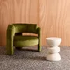 A green velvet armchair and a cream-colored side table are placed on a speckled floor in front of a wood-paneled wall. Produktbild-Vorschau