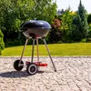 A black kettle-style grill with chrome legs and wheels sits on a stone patio in a sunny garden. Náhľad obrázku produktu