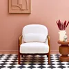 A white fluffy armchair with wooden legs is placed in a room with pink walls and a black and white checkered floor. A small side table with a vase of red flowers and a small statue is next to the chair. Produktbild-Vorschau