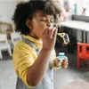 Kind in gelbem Hemd und Latzhose bläst Blasen aus einer kleinen blauen Flasche mit weißem Verschluss Náhled obrázku produktu
