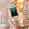 A close-up shot of a rectangular watch with a green dial and a gold mesh band, placed next to a decorative glass object. Produktbild-Vorschau