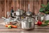 A collection of stainless steel pots and pans with glass lids are arranged on a wooden surface, with some vegetables and a bottle of wine in the background. Anteprima dell’immagine prodotto