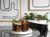 Two WoodWick candles are displayed on a marble tray with a small plant, in a home setting with abstract art in the background. Voir les images du produit