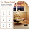A floor lamp with a woven shade is positioned next to a chair in a room with a window overlooking a city at dusk. Podgląd zdjęcia produktu