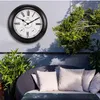A large outdoor clock with thermometer and hygrometer mounted on a gray wall next to lush green plants and a wicker sofa with white cushions. Produktbild-Vorschau