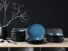 A table setting with a stack of blue and black plates and bowls, a vase with branches, and cutlery. Produktbild-Vorschau