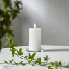 A white fluted LED candle with a flame sits on a clear glass coaster on a white surface. Green ivy leaves are in the foreground and to the left. Podgląd zdjęcia produktu