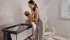 A woman is holding a baby next to a crib in a room. Produktbild-Vorschau