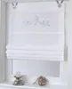 A white Roman blind with a silver crown motif is displayed in a window frame, with a candle holder and a decorative heart on the windowsill below. Produktbild-Vorschau
