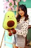 A person is holding a plush avocado toy in front of a backdrop with pink flowers and a ladder. Produktbild-Vorschau