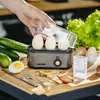 A hand is placing eggs into an egg cooker on a wooden table, surrounded by fresh vegetables and cutlery. Anteprima dell’immagine prodotto