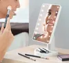 A woman is applying makeup using a makeup brush while looking into a lighted makeup mirror. Makeup brushes and lipsticks are on the table in front of the mirror. Produktbild-Vorschau