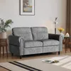 A gray sofa is placed in a living room setting with a plant, a lamp, and a table in the background. Produktbild-Vorschau