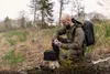 A man is kneeling in a forest setting, holding a small electronic device and looking at it. A black case is on a tree stump next to him, and he has a large backpack on. Produktbild-Vorschau