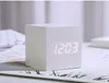 A white cube-shaped digital alarm clock with a bright LED display showing the time is placed on a desk with some papers and a keyboard in the background. Produktbild-Vorschau