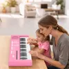A woman and a young girl are sitting at a table, with the girl playing a pink toy piano. The background shows a blurred living room setting. Produktbild-Vorschau