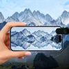 A hand holding a smartphone with a clip-on lens attachment, displaying a reflection of mountains on the phone screen, with a mountain landscape in the background. Voir les images du produit