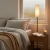 A cozy bedroom scene with a bed, pillows, a blanket, a bedside table with a book, candle, and mug, and a floor lamp. Náhľad obrázku produktu