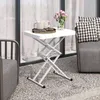 A white foldable side table with a scissor-like base is set up on a speckled outdoor patio, next to a gray chair with a black and white patterned cushion. Two mugs and a small tray with cookies are on the table, along with a vase of pink flowers. Produktbild-Vorschau