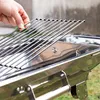 A hand is holding a barbecue grill rack over a stainless steel barbecue. Produktbild-Vorschau
